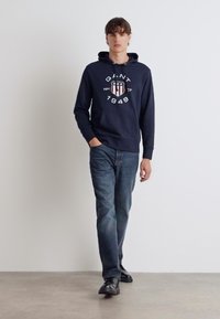 Sudadera azul marino con un logo en el frente, cordón y bolsillo frontal. Combinada con vaqueros azules medios y zapatos negros. Diseño minimalista.