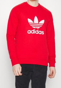 Röd sweatshirt med långa ärmar och rundad halsringning. Har en vit Adidas-logga med trebladig design och horisontella ränder.