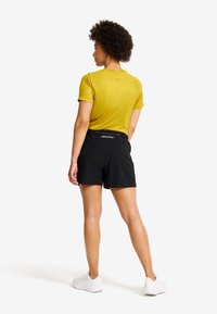 Gul kortärmad skjorta med texturerad yta, tillsammans med svarta shorts. Modellen har vita sneakers på sig, med en synlig logodetalj på shortsen.