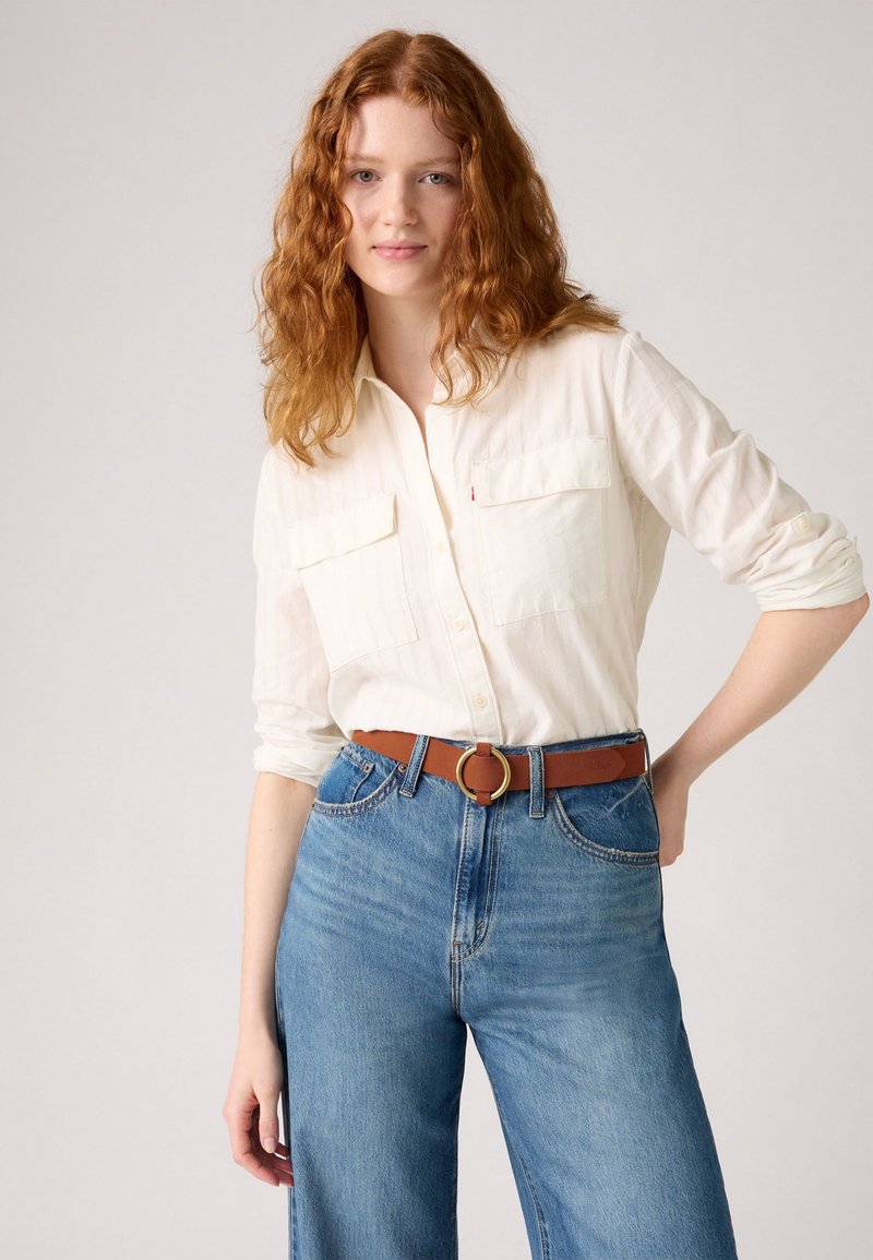 Femme aux cheveux roux portant une chemise blanche à boutons avec des manches retroussées, un jeans bleu taille haute et une ceinture marron avec une boucle en or ronde.