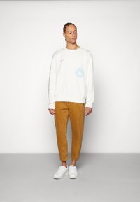 Pulover alb cu detalii grafice albastre și logo portocaliu. Pantaloni bej tapered dintr-un material texturat, asortați cu pantofi sport albi.