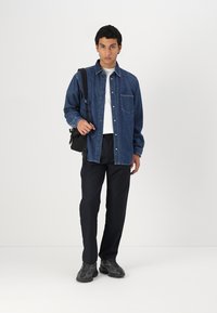 Camisa de mezclilla en azul oscuro con botones al frente y un solo bolsillo en el pecho, llevada sobre una camisa blanca. Pantalones negros y zapatillas completan el conjunto.