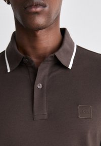 Braunes Poloshirt mit einem weißen Kragenrand, zwei Knöpfen und einem strukturierten Stoff. Hat ein kleines quadratisches Logo auf der linken Seite.