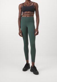 Dark green leggings with high waist and fitted design, paired with a black strappy sports bra. Model wearing black sneakers.