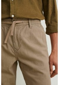 Pantalon jogger en lin marron clair avec une ceinture élastique et un cordon de serrage. Poche avant visible, tissu avec une finition douce et texturée.