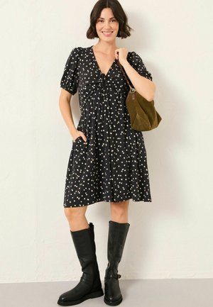 Schwarzes, kurzärmeliges Kleid mit weißen Polka-Dots, an der Taille gerafft, mit Taschen; kombiniert mit hohen schwarzen Lederstiefeln und einer olivgrünen Tasche.