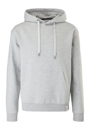 Person iført en lysegrå hættetrøje, blå jeans og sorte snørebåndsstøvler, står imod en ensfarvet baggrund med hænderne i lommerne.