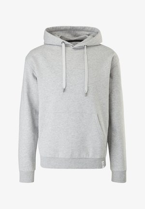 Personne portant un sweat à capuche gris clair, un jean bleu et des bottes noires à lacets, debout devant un fond uni, les mains dans les poches.