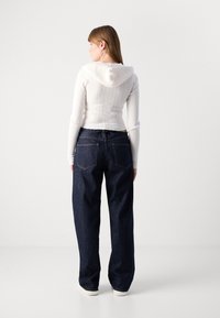 Maglione bianco a coste con cappuccio, abbinato a jeans di jeans scuri a vestibilità ampia. I jeans hanno tasche posteriori e cuciture a contrasto.