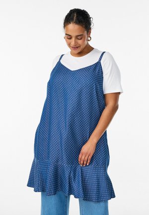 Moteris dėvi baltą trumpomis rankovėmis palaidinę po mėlyna su taškeliais berankove suknele su raukšlėtu apdaila, derina su šviesiai mėlynomis džinsais, žvelgia žemyn.