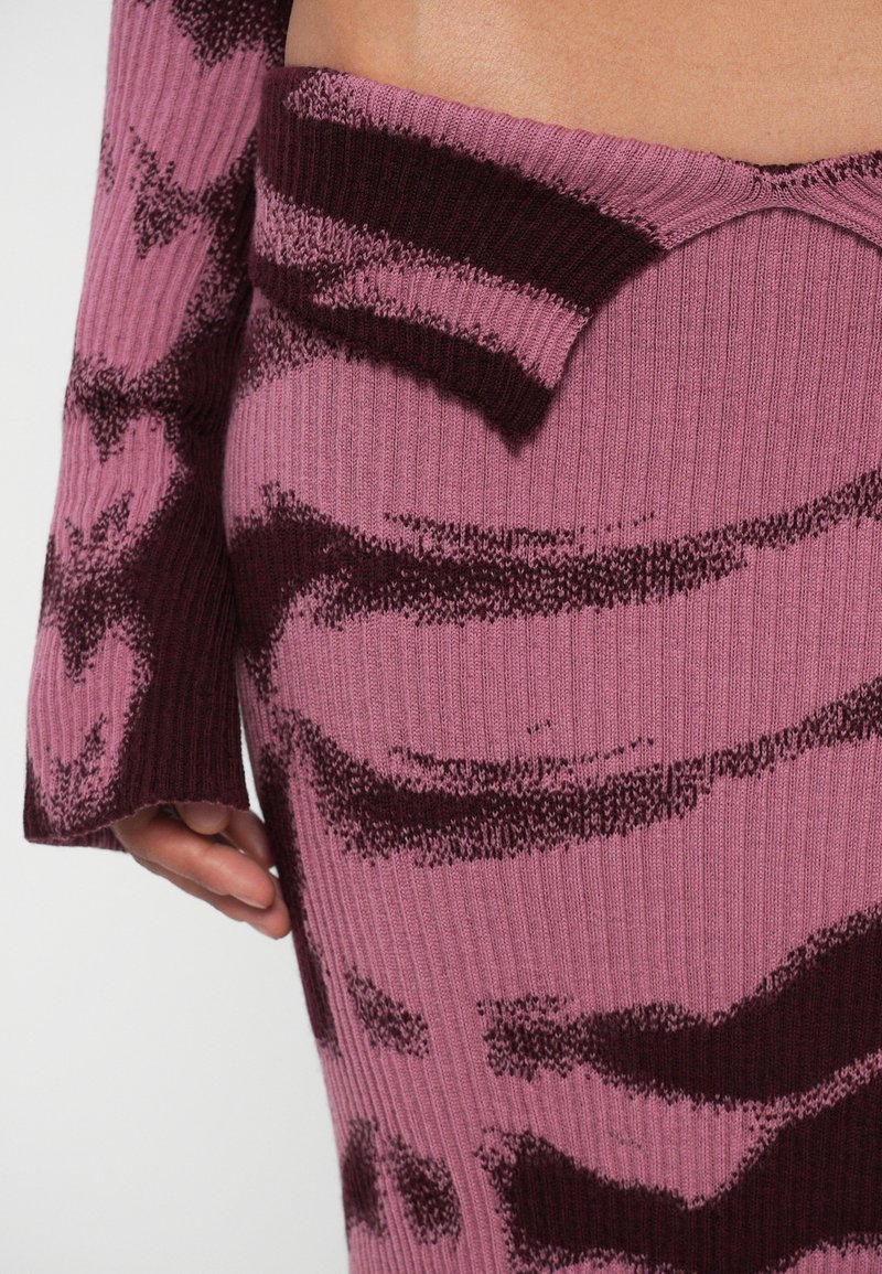 Ribbat stickad klänning i rosa och vinröd med vågmönster, med en asymmetrisk halsringning och långa ärmar. Mjuk, texturerad tyg.