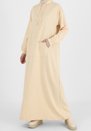 Femme portant une longue robe à capuche beige avec une poche avant et une capuche, associée à des chaussures à plateforme beige assorties.