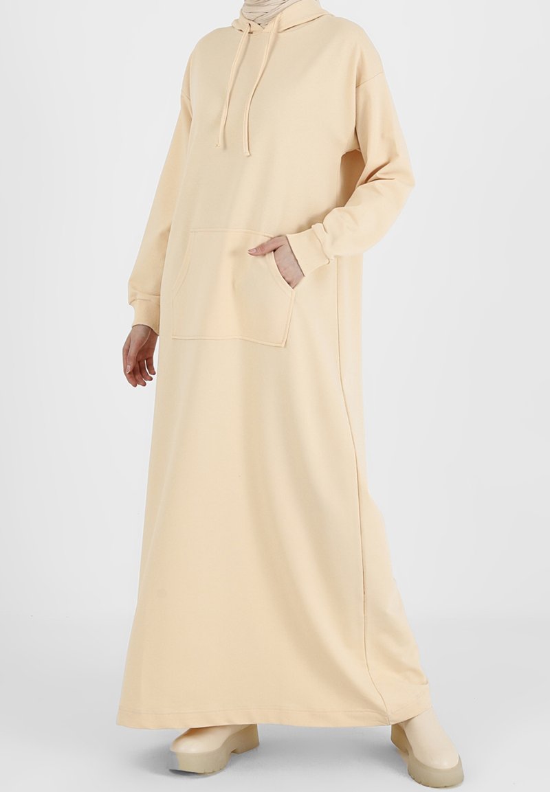 Femme portant une longue robe à capuche beige avec une poche avant et une capuche, associée à des chaussures à plateforme beige assorties.