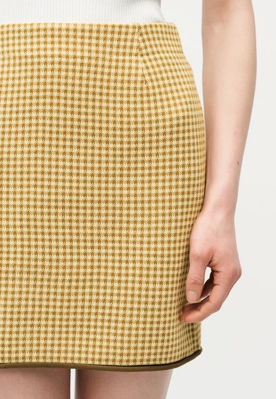 Φούστα με κίτρινο και λευκό σχέδιο ψαροκόκκαλο και σκούρο περίγραμμα, φορεμένη με λευκό πλεκτό τοπ, που δείχνει το πλάι του χεριού και της μέσης ενός ατόμου.