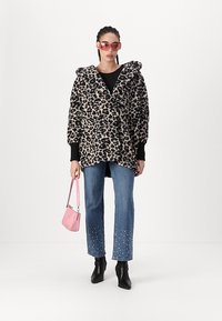 Leopardprint oversized jas met een capuchon, zwarte lange mouwen, gecombineerd met lichtblauwe jeans versierd met witte kralen en zwarte enkellaarzen.