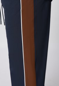 Μπλε παντελόνι ναυτικού σκούρου με κάθετη καφέ ρίγα περιτριγυρισμένη από λεπτές λευκές γραμμές κατά μήκος του ποδιού, με μαύρα τελειώματα κορδονιών.