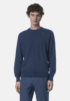 Młody mężczyzna z kręconymi włosami, ubrany w gładki granatowy sweter z długim rękawem oraz pasujące granatowe spodnie, stojący na białym tle.