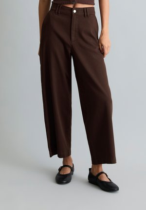 Femme portant un pantalon large taille haute marron foncé et des ballerines noires Mary Jane, debout devant un fond gris uni.