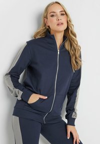 Felpa con zip colore navy, dotata di pannelli laterali grigi, maniche lunghe e tasche frontali; realizzata in tessuto morbido con colletto e polsini a coste.