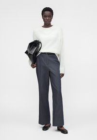 Femme portant un pull blanc et un pantalon gris à jambes larges, tenant un sac en cuir noir sous le bras, debout devant un fond uni.