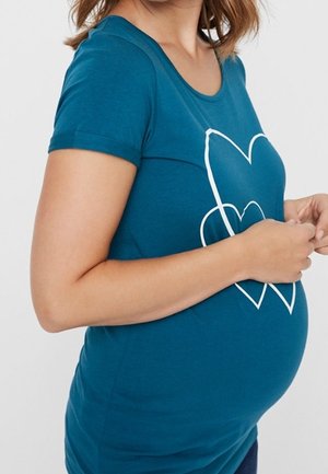 Zwangere vrouw in een groenblauw shirt met witte hartcontouren, die zachtjes haar buik vasthoudt, zichtbaar van schouders tot heupen.