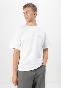 En blond mann som har på seg en hvit T-skjorte med kort erm og liten tekst på brystet, samt grå jeans. Han står med den ene hånden i lommen og ser til siden.