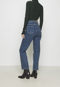 Jeans azules de talle alto con corte de pierna recta, dos bolsillos traseros y una etiqueta de cuero en la cintura. Combinados con una camiseta de manga larga verde oscuro y botas negras con tacón.