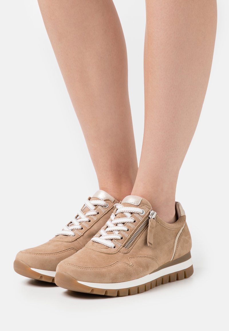 Beige suede sneakers with white laces, silver accents, side zipper, and textured rubber soles, featuring a cushioned heel for support.