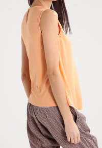 Persikofärgad ärmlös topp med en lös silhuett, med en nyckelhålsdetalj på ryggen och mjukt, slätt tyg. Matchad med mönstrade, vida byxor.