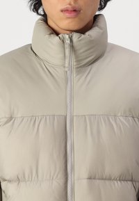 Puffer jacket in light beige with a high collar and full zipper. Features a quilted design with a smooth, shiny texture.