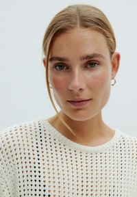 Weißer Strickpullover mit offenem Muster und schwarzen Akzenten. Nahaufnahme einer Person mit natürlichem Make-up und hellbraunen Haaren.