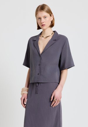 Frau mit kurzen blonden Haaren, die ein locker sitzendes, violettes Hemd mit Knöpfen und einen passenden Rock trägt, dazu Goldschmuck und transparente Armreifen als Accessoires.