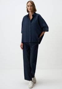 Navyblaues, oversized Hemd mit Knöpfen und Kragen, hochgerollten Ärmeln, kombiniert mit dunklen Jeans im weit geschnittenen Stil und weißen Sneakers.