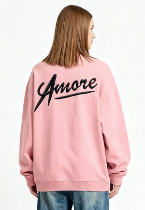 Persoon met steil bruin haar die een losse roze trui draagt met "Amore" in grote zwarte letters op de rug, gecombineerd met een blauwe spijkerbroek.