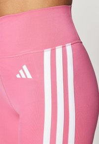 Roze high-waisted leggings met een gladde textuur, voorzien van drie verticale witte strepen en een wit logo op de zijkant.
