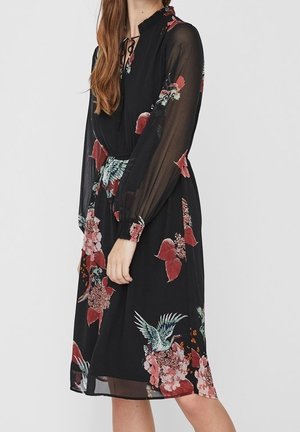 Femme portant une robe midi noire à manches longues transparentes avec des imprimés floraux et d’oiseaux dans des tons rouges et verts.