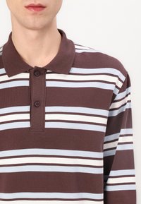 Camisa polo de mangas longas castanha com riscas, apresentando um colarinho clássico, dois buttons e riscas alternadas em azul claro e branco. Tecido com textura suave.