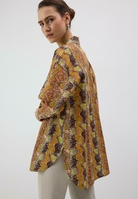 Chemise à manches longues avec un motif serpent coloré en orange, jaune et marron, dotée d'un ourlet arrondi et de fentes latérales pour une coupe décontractée.