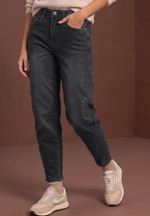 Le mannequin porte un jean noir clouté, un pull rose pâle et des baskets beiges, et pose avec une main dans la poche.