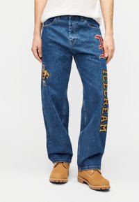 Τζιν παντελόνι σε μεσαίο μπλε, άνετη εφαρμογή, με κεντημένη αρκούδα και έντονο κείμενο "ICECREAM" σε κίτρινο. Ομαλή υφή με ορατές ραφές.