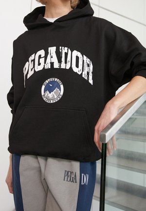 Personne portant un sweat à capuche Pegador noir avec un logo de montagne et le texte "No love lost, no love found", assorti à un pantalon bicolore avec la marque Pegador.