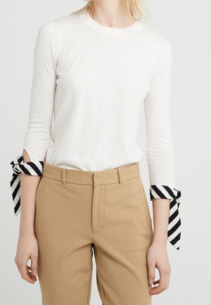 Kobieta w białej koszuli z długim rękawem, z czarno-białymi paskami przy mankietach i wysokim stanem beżowych spodni.