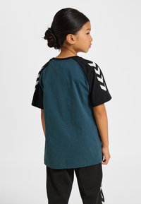 Barn-T-shirt med turkos kropp, svarta ärmar och vita chevron-detaljer på axlarna; bomullstyg med avslappnad passform.