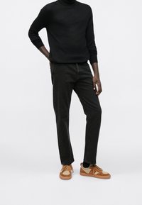 Maglione a dolcevita nero, pantaloni verde scuro e sneakers bicolore marroni con dettagli testurizzati, in piedi contro uno sfondo bianco neutro.