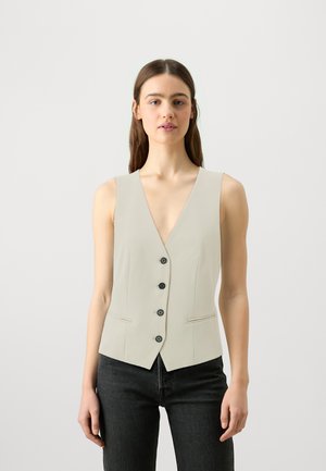 Beige ermeløs vest med dyp V-hals, fem svarte knapper foran og sidelommer. Båret over mørke jeans. Glatt stofftekstur.