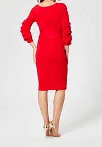 Rotes, figurbetontes Kleid mit langen Ärmeln, tailliertem Design und knielangem Saum. Getragen mit goldenen High Heels, die eine glatte Textur zeigen.