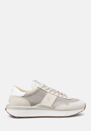 Sneakers featuring a beige mesh upper with suede accents, a white rubber sole, and a textured gray panel. Laces and logo in matching tones.