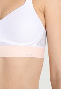 Hvit bh med teksturert mesh-design, myk rosa kant og subtil merkevare. Har runde kopper og justerbare stropper for komfort.