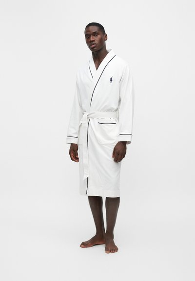 Homme debout pieds nus portant une robe blanche avec des bordures noires et une ceinture nouée sur un fond blanc uni.