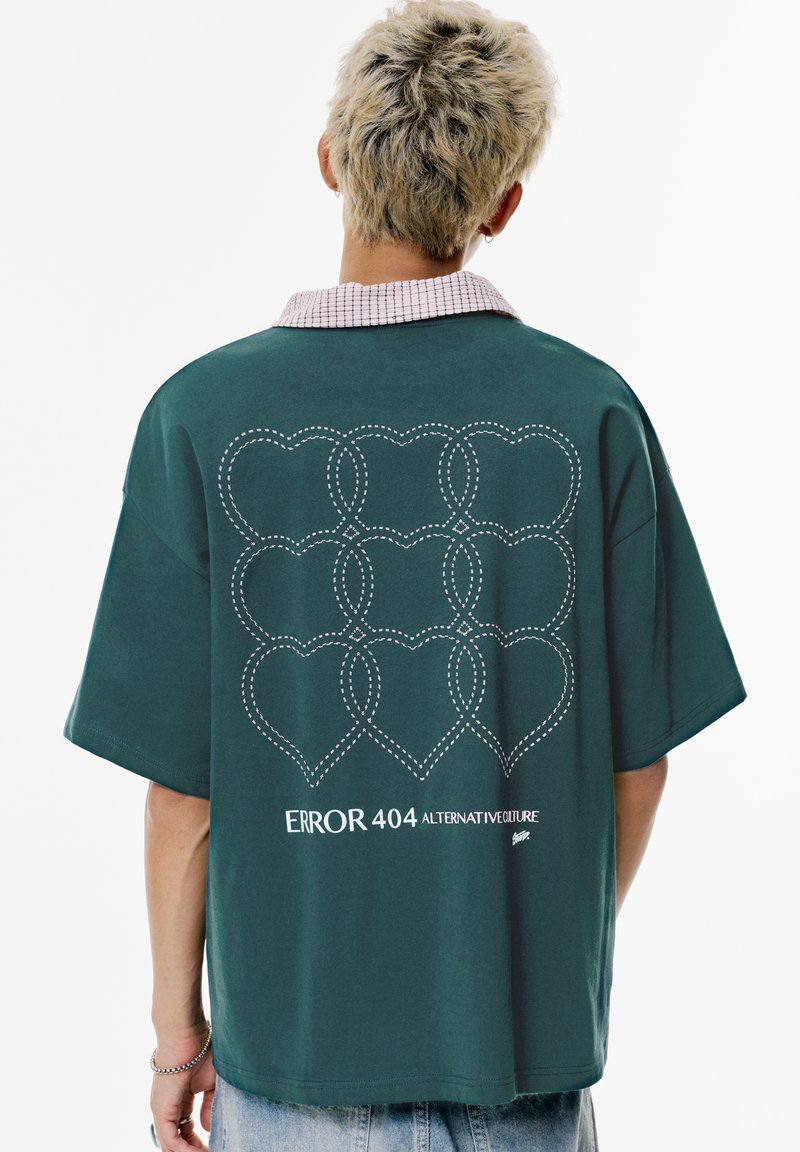 Oversized groene katoenen t-shirt met korte mouwen, voorzien van een wit hartpatroon en de tekst "ERROR 404 ALTERNATIVECULTURE" op de achterkant.
